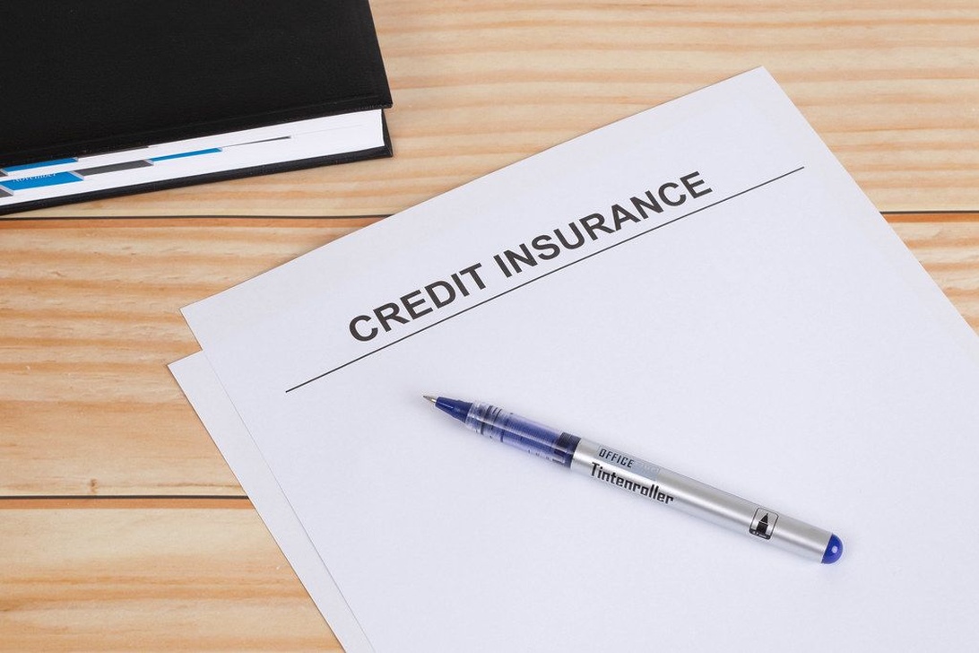 A formal document titled 'CREDIT INSURANCE' with a blue pen placed on top, positioned on a wooden desk surface with a black book visible in the corner. This directly represents credit insurance documentation that would be used in the credit insurance business.